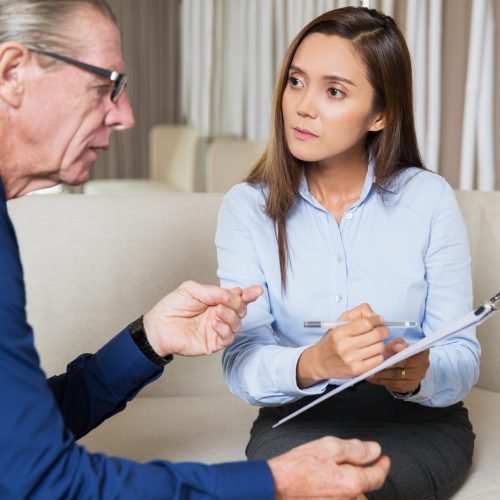 Serious young woman sitting on sofa, writing and showing questionnaire to senior man who is answering and sitting sideways to camera